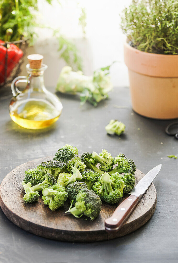 Broccoli on wooden cutting board with kitchen knife at grey kitchen table with herbs, oil and ingredients at background with natural lights