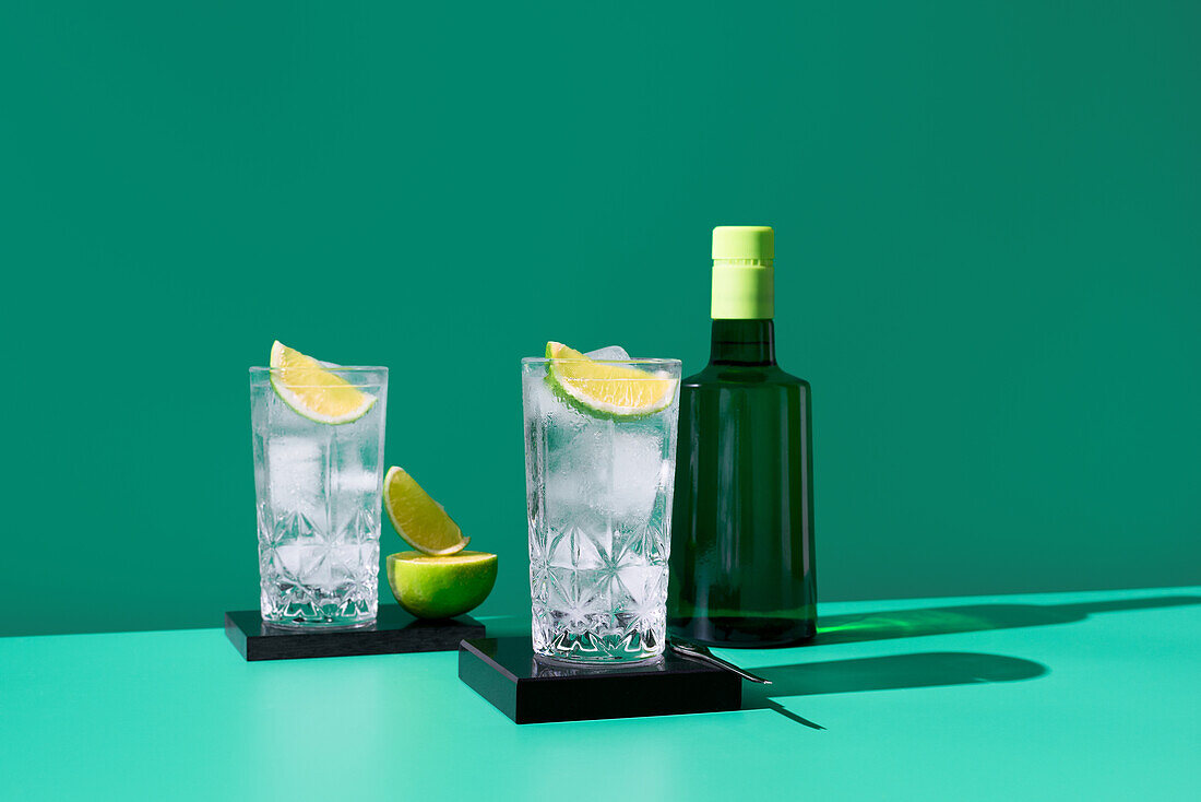Eine künstlerische Komposition mit zwei Gläsern mit Gin Tonic und Limettenspalten sowie einer dunklen Flasche Whiskey vor einem leuchtend grünen Hintergrund