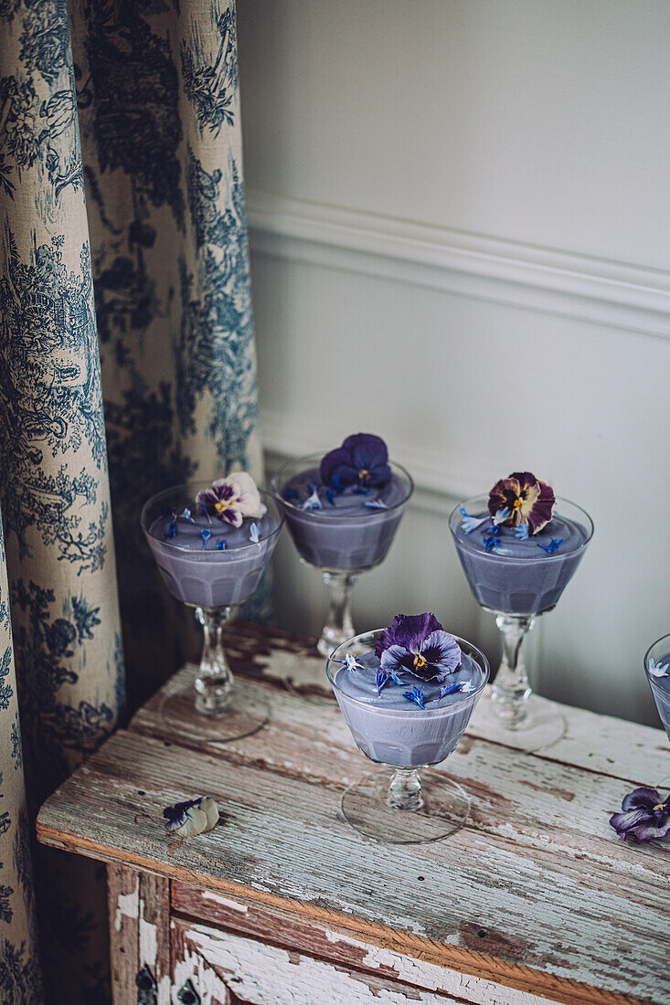 Verschiedene Gläser mit schmackhaftem, blauem Spirulina-Pudding, dekoriert mit Blumen, auf einem schäbigen Holzschrank