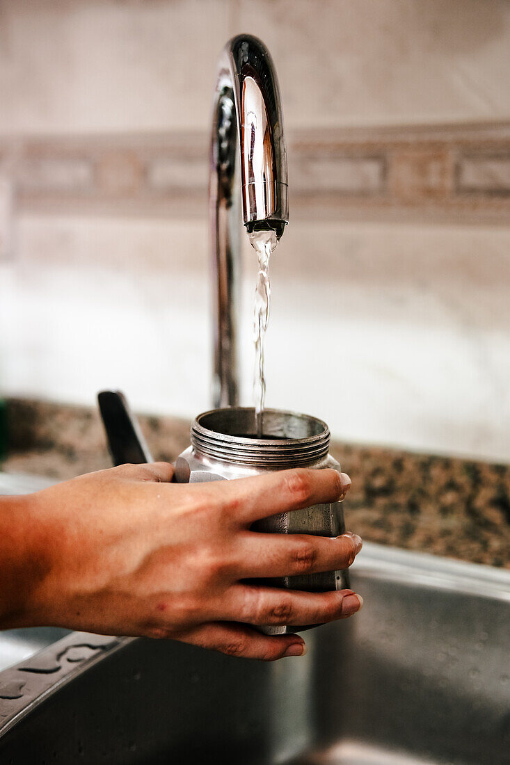 Nahaufnahme einer anonymen Hand, die eine italienische Mokkakanne mit Wasser aus einem Küchenhahn füllt, um die ersten Schritte der Zubereitung von traditionellem, selbst gebrühtem Kaffee hervorzuheben