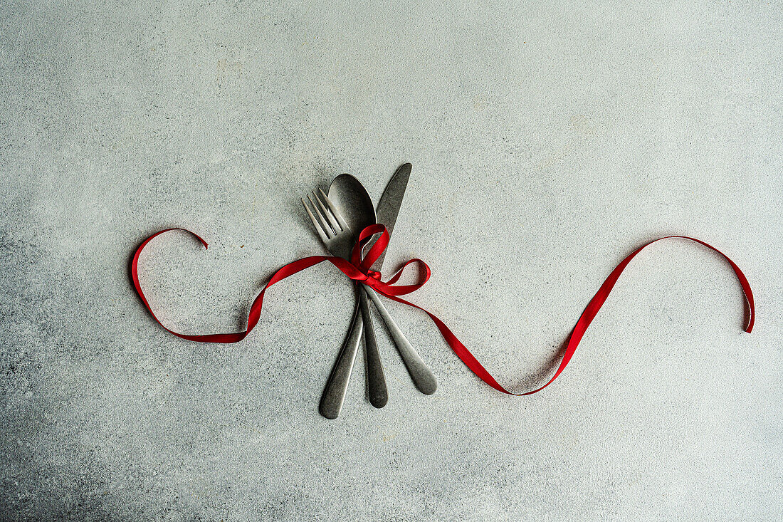 Top view of vintage cutlery set placed on gray surface with red ribbon in light kitchen