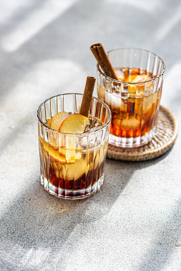 From above of fresh apple cider cocktails garnished with cinnamon sticks, star anise, and apple slices in a crystal glass filled with ice cubes on a textured background