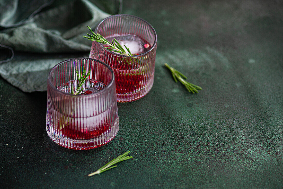 Two glasses of gin tonic cocktail with pomegranate seeds and a sprig of rosemary, on a dark green textured surface with a single rosemary sprig beside