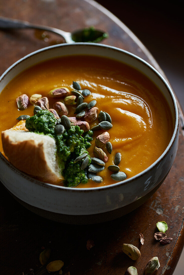 A warm bowl of creamy pumpkin soup garnished with pumpkin seeds, pistachios, and vibrant green pesto, served on a rustic wooden surface