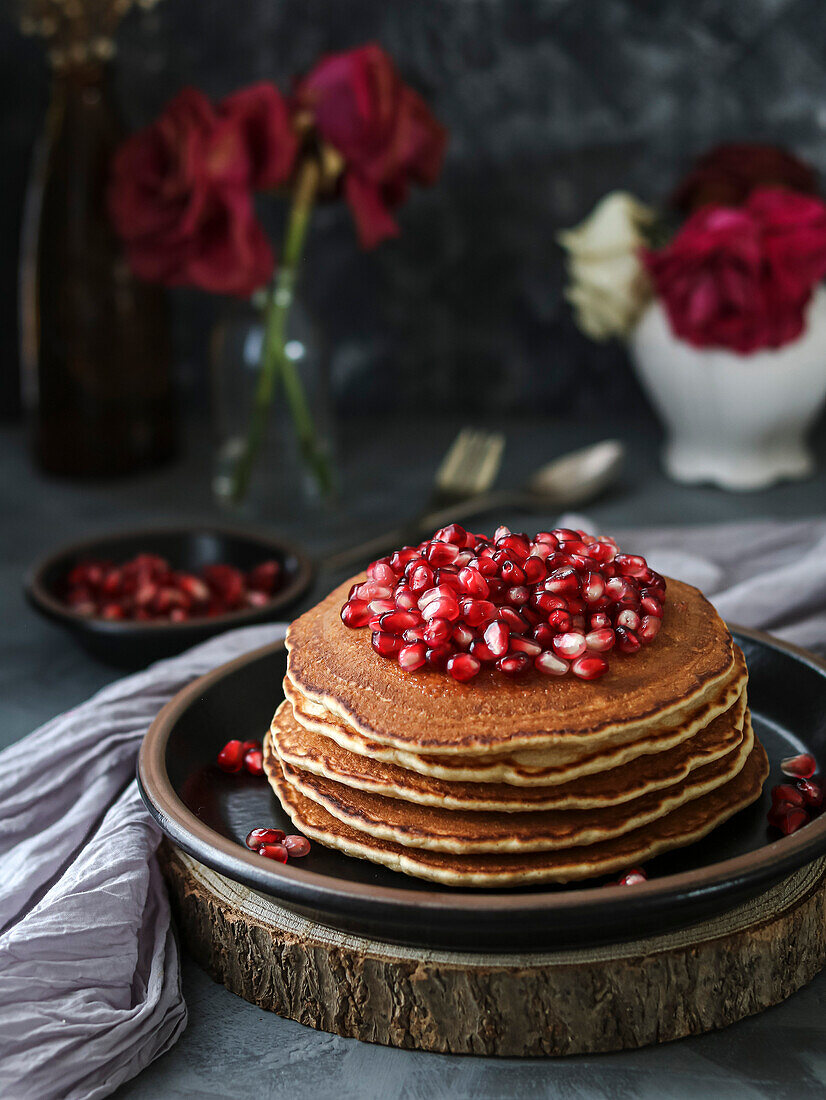 Stapel Pfannkuchen mit Granatapfel und roten Blumen auf einem schwarzen Teller und einem dunklen Hintergrund