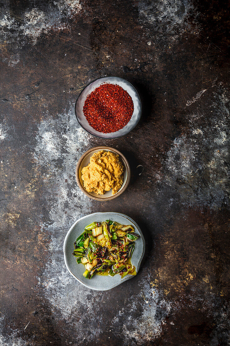 Three small bowls with chilli flakes, miso paste and charred shallots on a dark background