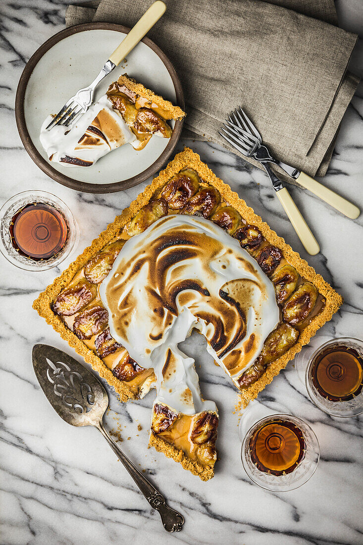 Banana Cream tart with toasted marshmallow topping. One slice cut, on marble surface with rum in vintage glasses