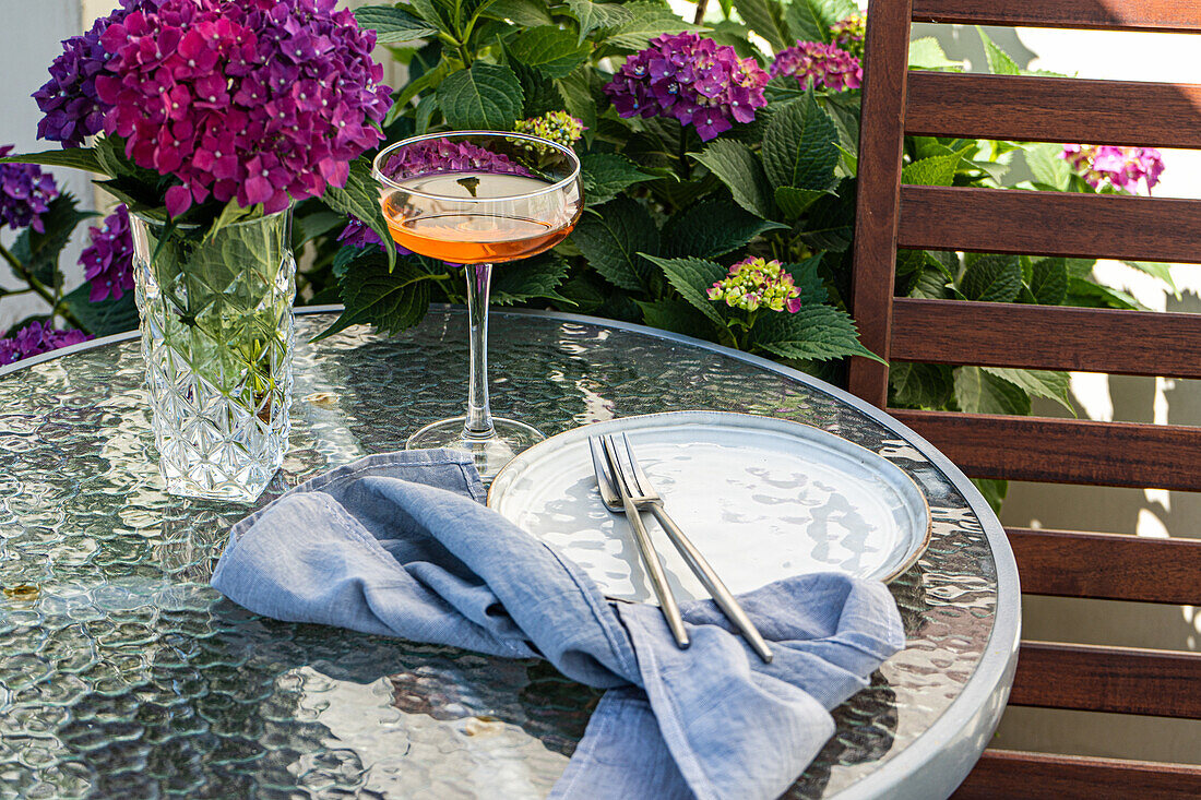 Vase mit lila Hortensien steht auf dem Tisch neben einem Glas mit Getränk und einem Keramikteller im Garten