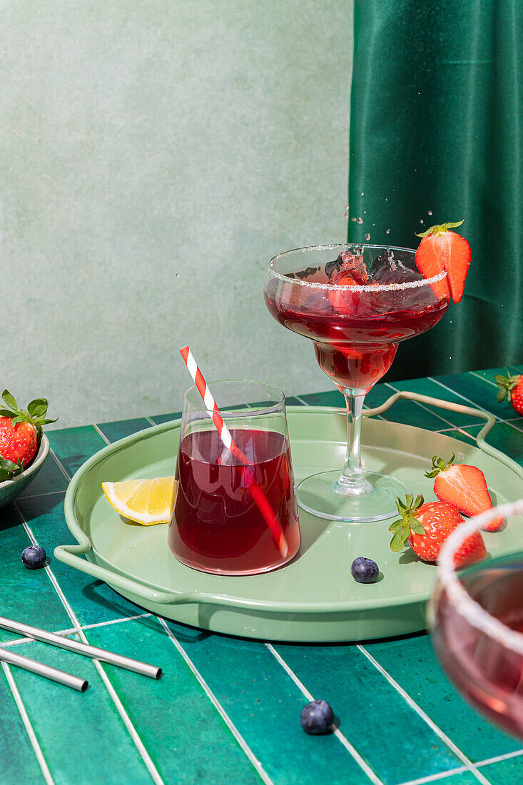 Gläser mit erfrischenden roten alkoholischen Cocktails mit Erdbeeren und Strohhalm, serviert auf einem Tablett mit Beeren und Zitronenscheibe auf dem Tisch