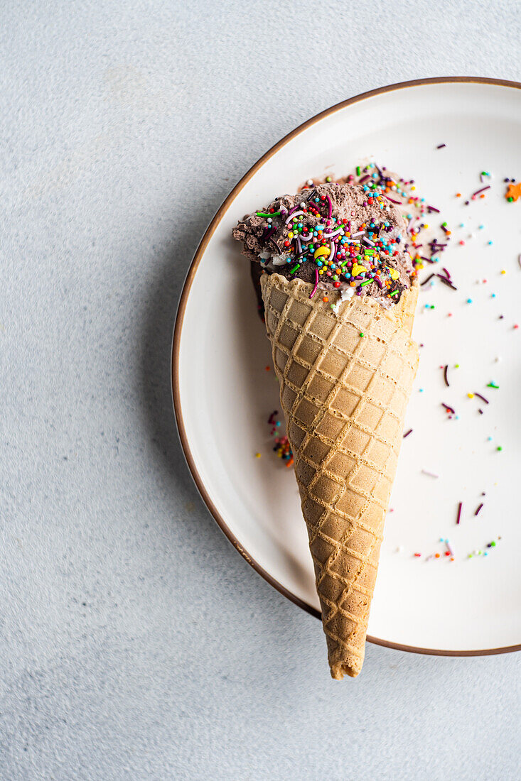 Top view of a waffle cone with coffee and chocolate ice cream with multicolored sugar sprinkles on a ceramic plate on a concrete background