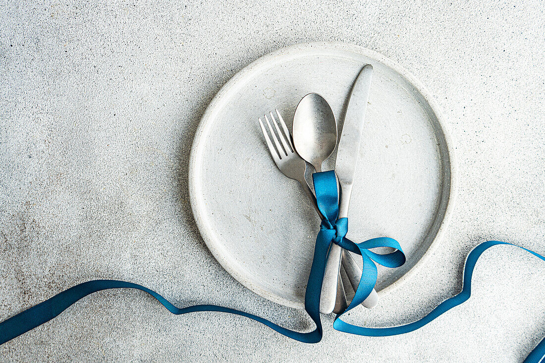 Top view of minimalist yet stylish Easter table setting, with cutlery tied together with blue ribbon on concrete plate, placed on gray table