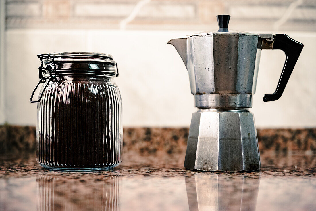 A ribbed glass jar with a metal clamp lid stands next to a vintage silver moka pot on a reflective granite countertop