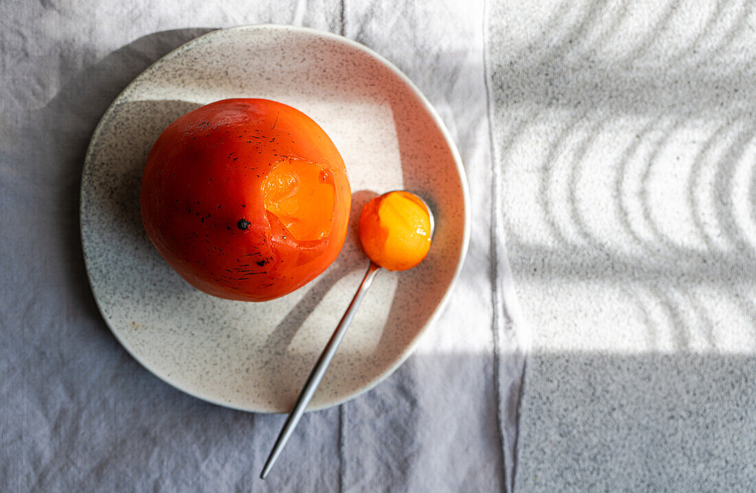 A vibrant, ripe persimmon rests on a ceramic plate beside a spoon, with dramatic shadows accentuating its curves.