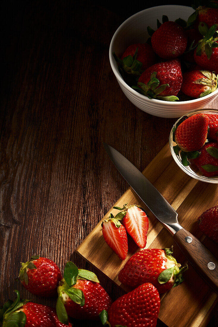 From above of fresh strawberries and knife on cutting board with cut juicy slices placed on wooden surface