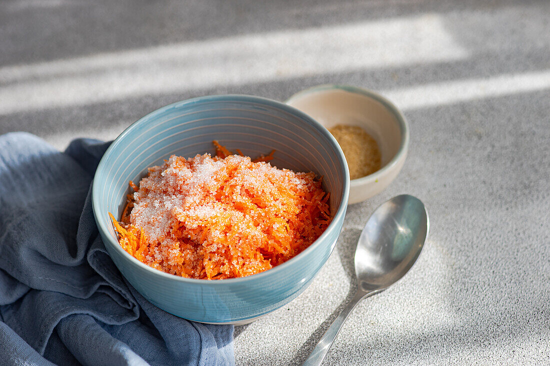 High angle of close-up of a light blue bowl containing freshly grated carrot sprinkled with sugar, placed next to a gray cloth, a small beige bowl with brown sugar, and a metallic spoon, all set against a textured gray background with sunlight casting soft shadows