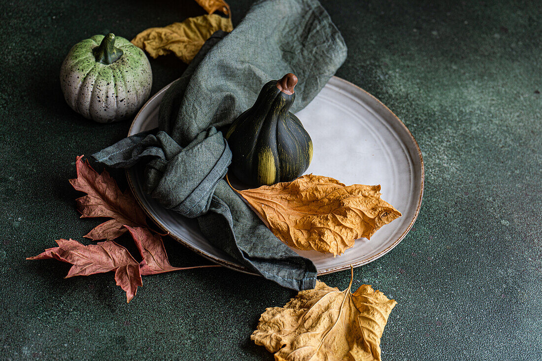 High angle of autumnal table setting with napkin, leaves and pumpkins placed on ceramic plate against dark surface