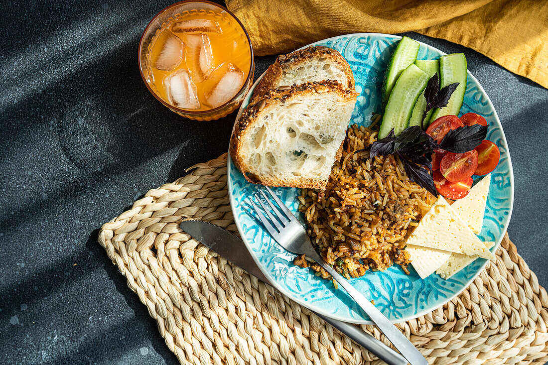 Draufsicht auf fünf Zutaten Mittagessen Set besteht aus hausgemachten Ziegenkäse, frische Gurken und Tomaten mit Blättern von Basilikum Kraut, zusätzlich zu gebratenem Reis mit Fleisch mit Scheiben von gesunden Sauerteig Brot serviert mit Glas Karotte-Apfel-Saft gegen grauen Hintergrund