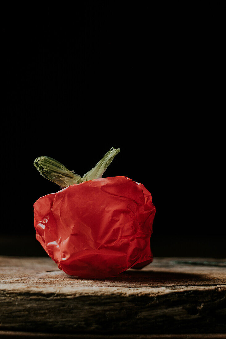 Eine rote Plastiktüte, die so geformt ist, dass sie einem Gemüse oder einer Frucht auf einer hölzernen Oberfläche vor einem schwarzen Hintergrund ähnelt
