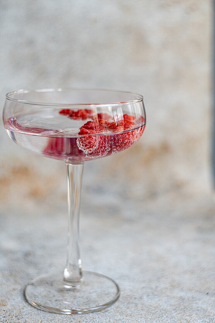 Transparenter glatter Glasbecher gefüllt mit erfrischendem Cocktailsekt mit reifen frischen Himbeeren vor unscharfem Hintergrund