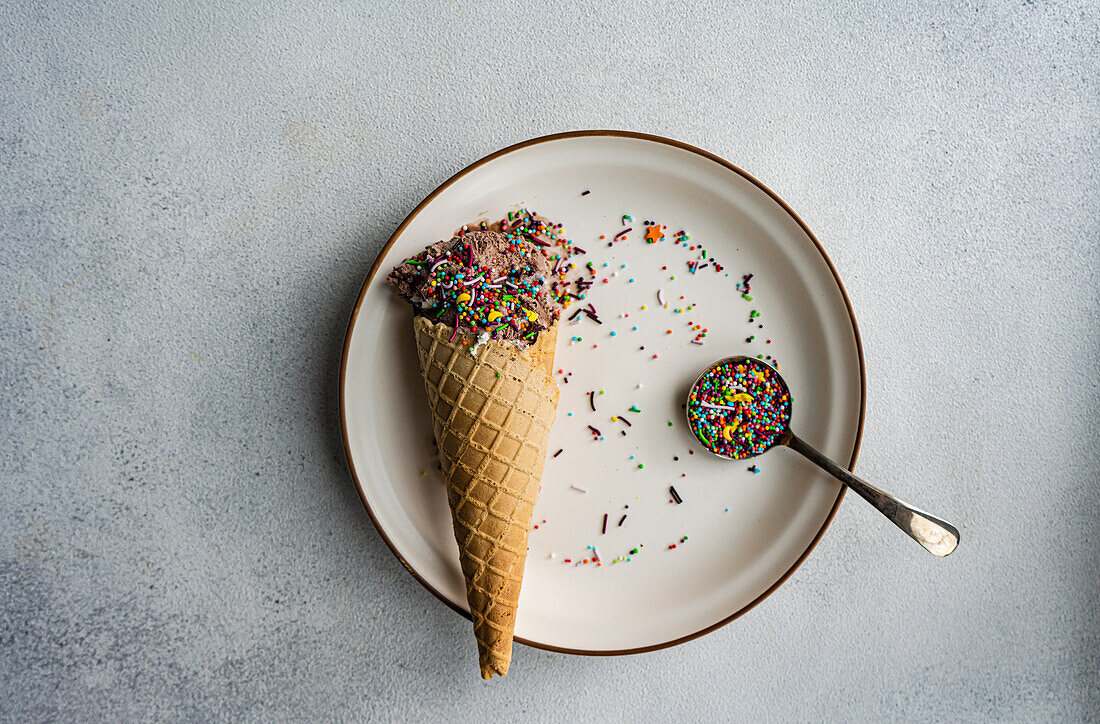Close up of a waffle cone with coffee and chocolate ice cream with a spoonful of multicolored sugar placed on a ceramic plate on a concrete background