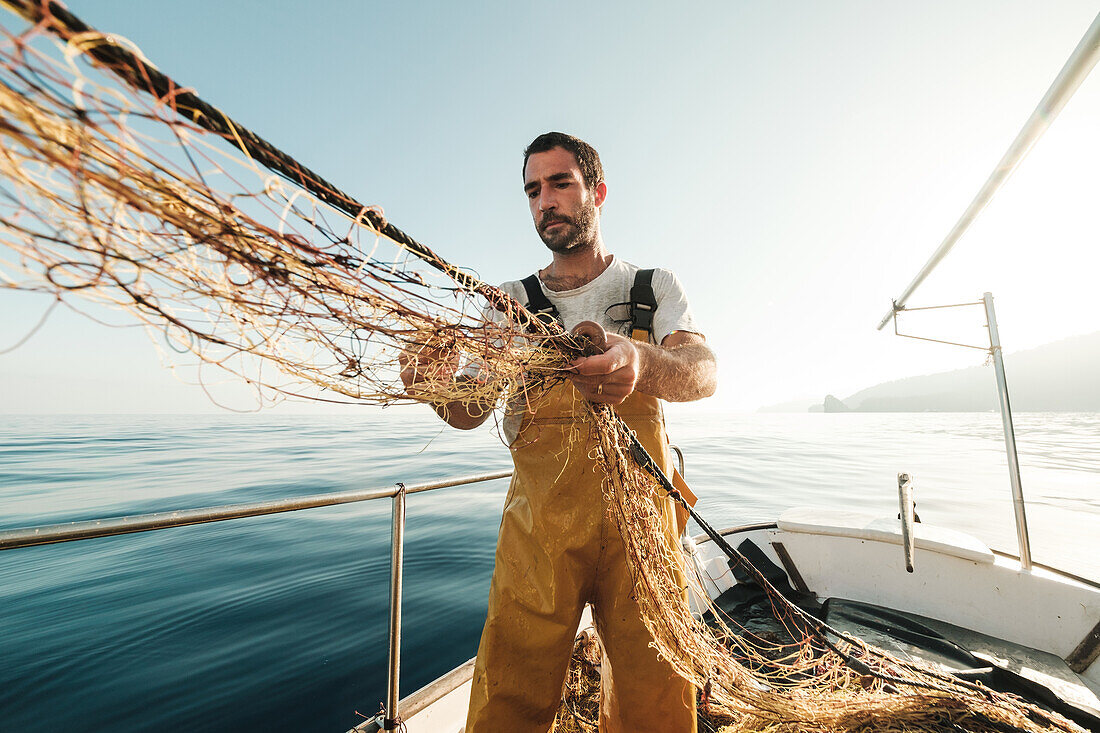 From below focused bearded male fisher in uniform seiner hunting fish with net while working on schooner in Soller near Balearic Island of Mallorca