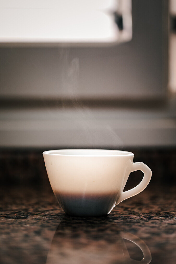 Eine dampfende weiße Kaffeetasse steht elegant auf einer Granitarbeitsplatte, die von oben mit sanftem Umgebungslicht beleuchtet wird und ein Gefühl von Ruhe und Morgenritual ausstrahlt