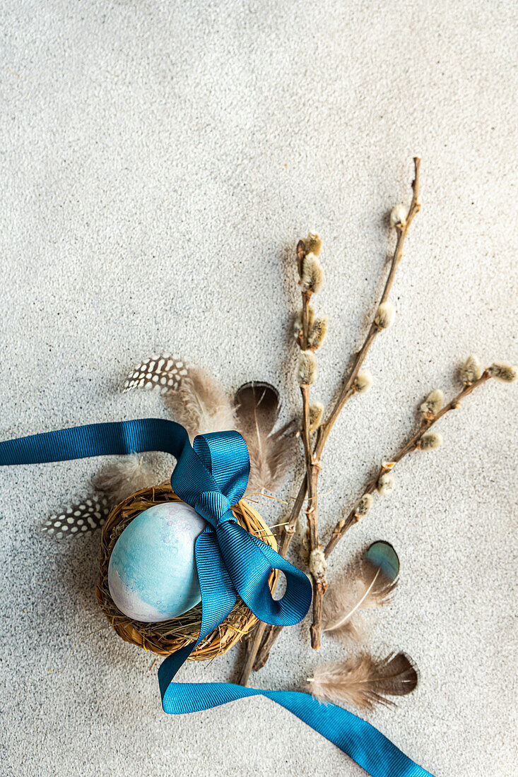From above of elegant Easter flatlay featuring a blue egg in a nest tied with a ribbon, accompanied by pussy willow branches and feathers on a textured surface