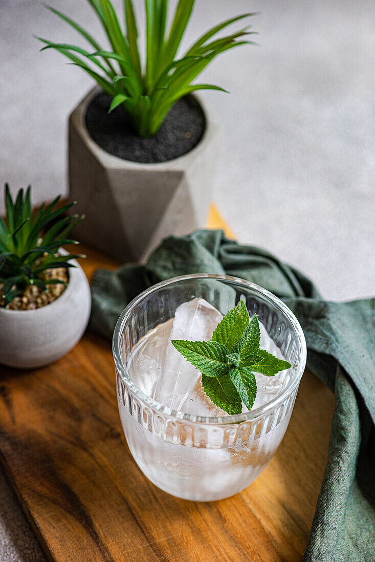 Blick von oben auf ein Glas Mineralwasser mit Eiswürfeln und frischen Minzblättern auf einem Holztablett mit Topfpflanzen und Serviette vor grauem Hintergrund