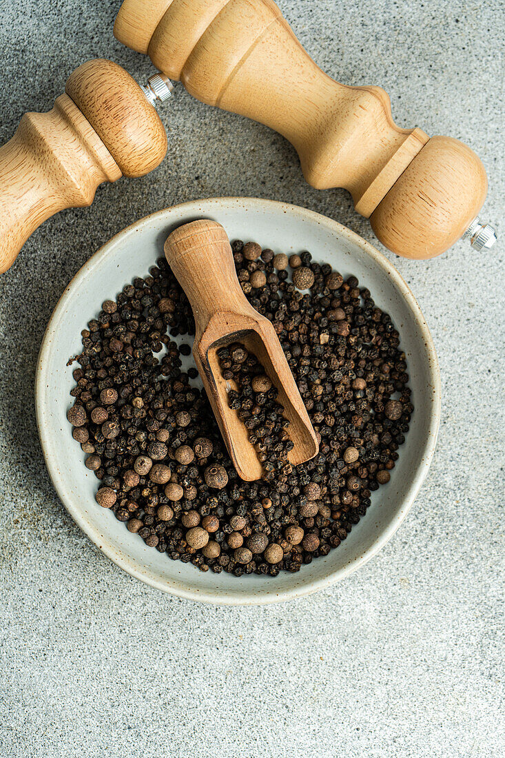 Top view of bowl filled with natural aromatic Black pepper in grain with wooden scoop on grey concrete background