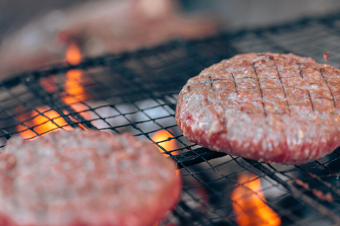 Beef burgers cook on a grill, with open flames licking the edges, capturing the essence of a summer barbecue