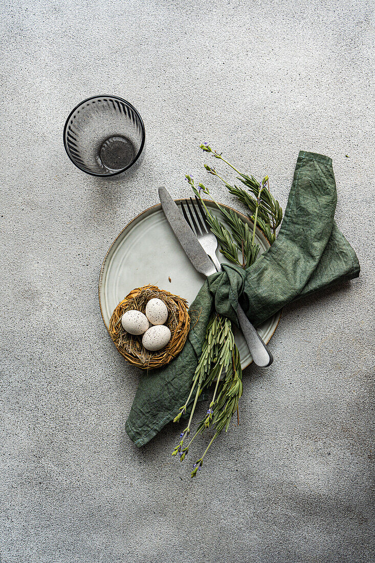 A top-view image featuring an elegant table setting with a rustic feel, including a linen napkin, ceramic plate, cutlery, a nest with eggs, and fresh greenery