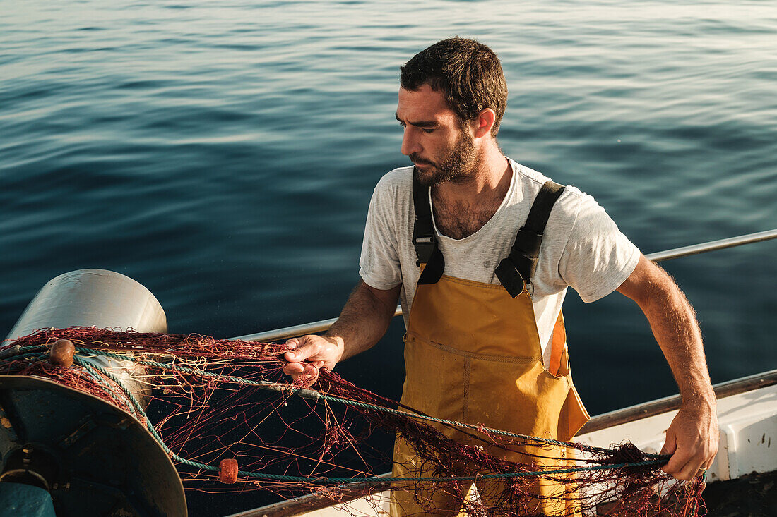 Von oben fokussierter bärtiger männlicher Fischer in Uniform, der mit einem Netz auf Fischfang geht, während er auf einem Schoner in Soller nahe der Baleareninsel Mallorca arbeitet