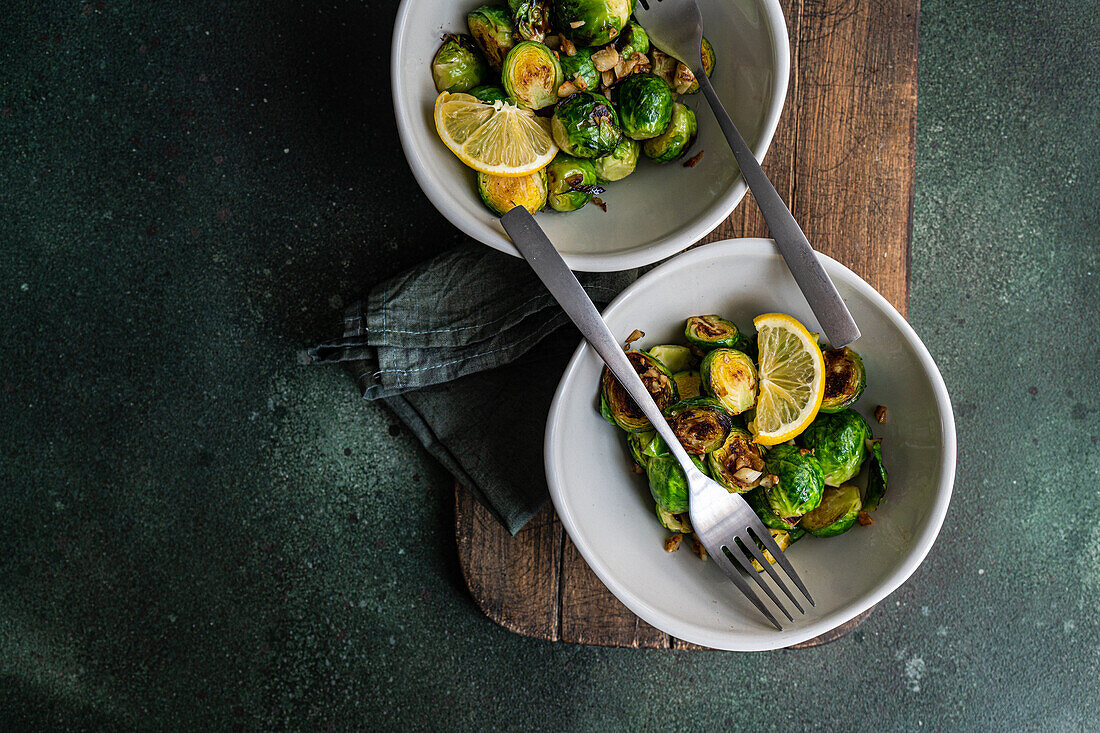 Top view of bowl of grilled Brussels sprouts seasoned with garlic and spices, garnished with a slice of lemon, served on a wooden board with a fork