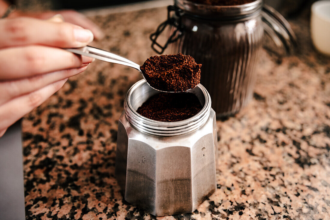 Close-up of anonymous person hand transferring freshly ground coffee from a jar to a stainless steel stovetop espresso maker set on a speckled countertop
