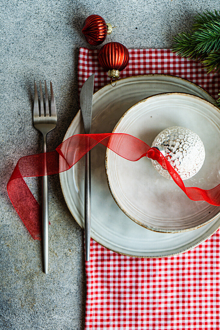 From above of plates with Christmas ball decorated with ribbon and fork and knife placed on gray table