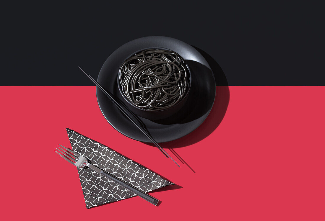 Draufsicht auf schwarze Spaghetti in einer Schüssel mit Stäbchen auf schwarzem und rotem Hintergrund neben Serviette und Gabel