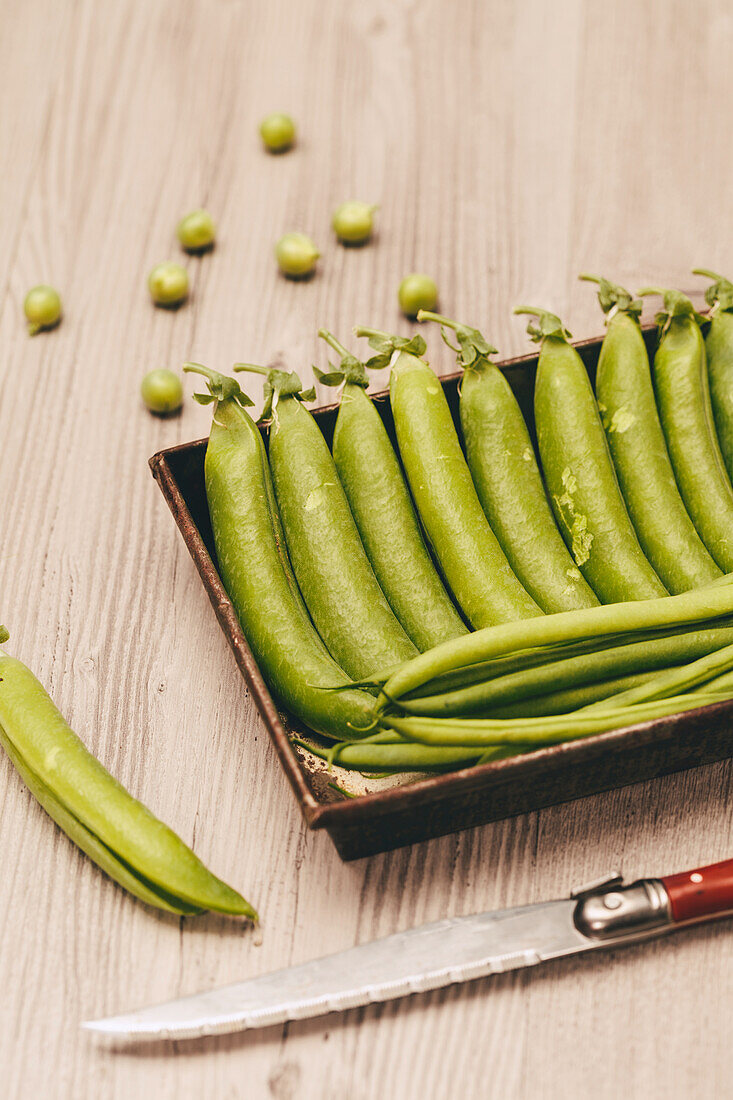 Ripe green peas arranged in a brown rustic tray next to a pocket knife on a wooden tabletop, with scattered peas around.