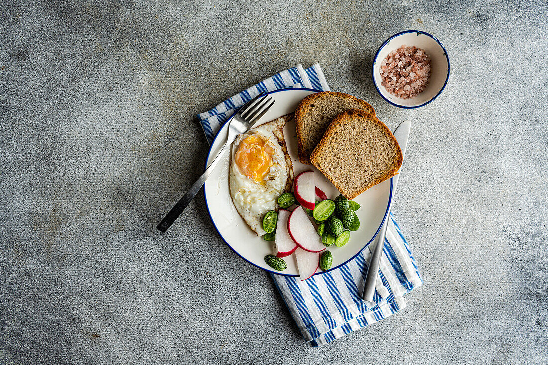 Draufsicht auf eine gesunde Mittagsschüssel mit Brotscheiben, Spiegelei, frischer Gurke, Rettich und Tomate auf einem blau-weiß gestreiften Tuch, daneben eine kleine Schale mit rosa Salz, vor einem grau strukturierten Hintergrund
