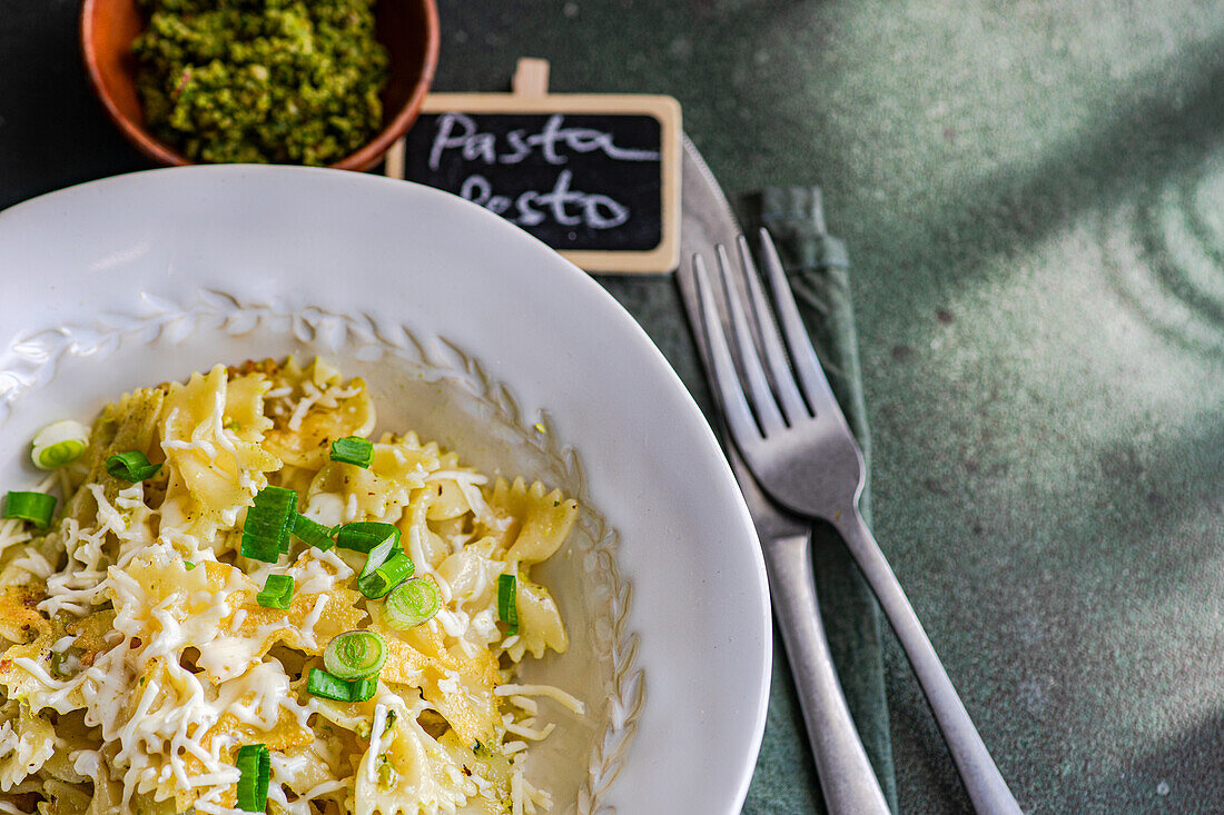 A plate of appetizing pesto Farfalle pasta sprinkled with cheese and herbs, with a fork on the side, and a chalkboard sign.