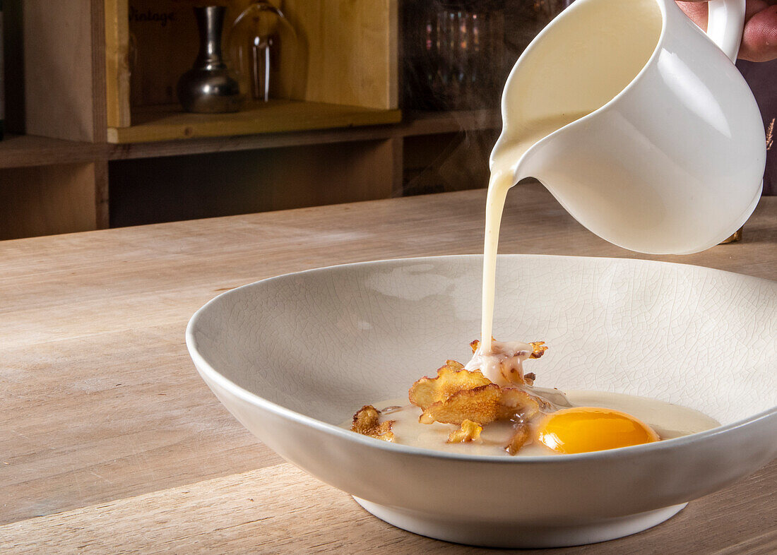 Closeup of creamy sauce pouring on top of fried strips with raw egg yolk in plate on wooden table