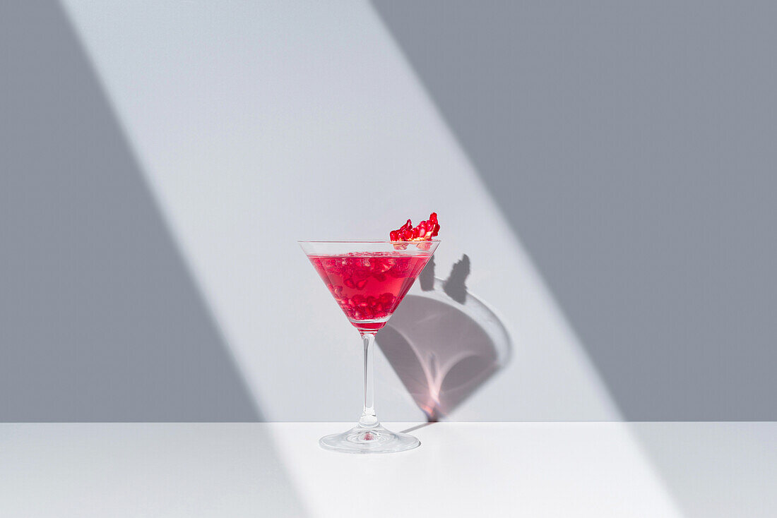 Mit rotem Granatapfelcocktail gefülltes Glas mit Granatapfelkernen auf einer spiegelnden Oberfläche unter einem weichen Lichtstrahl, der symmetrische Schatten erzeugt