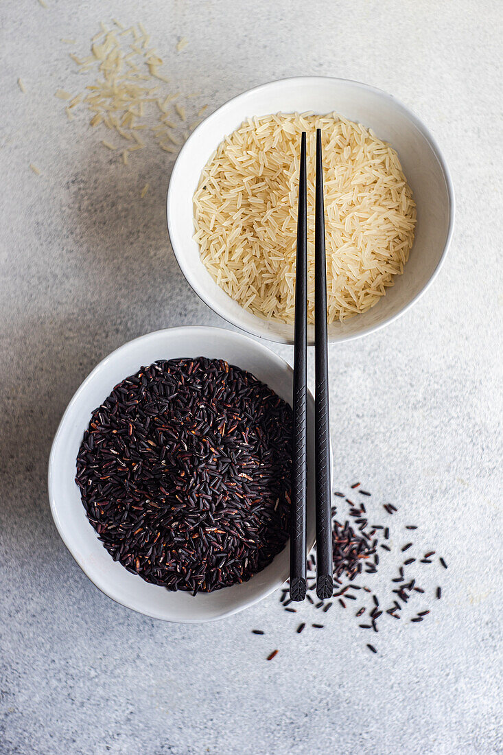 Top view of Raw wild black rice and peeled white rice in the bowls with chopsticks against white surface