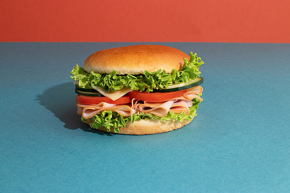 Leckeres Sandwich mit Schinken, Tomaten, Gurken- und Käsescheiben und frischem Salat auf blauem und orangem Hintergrund