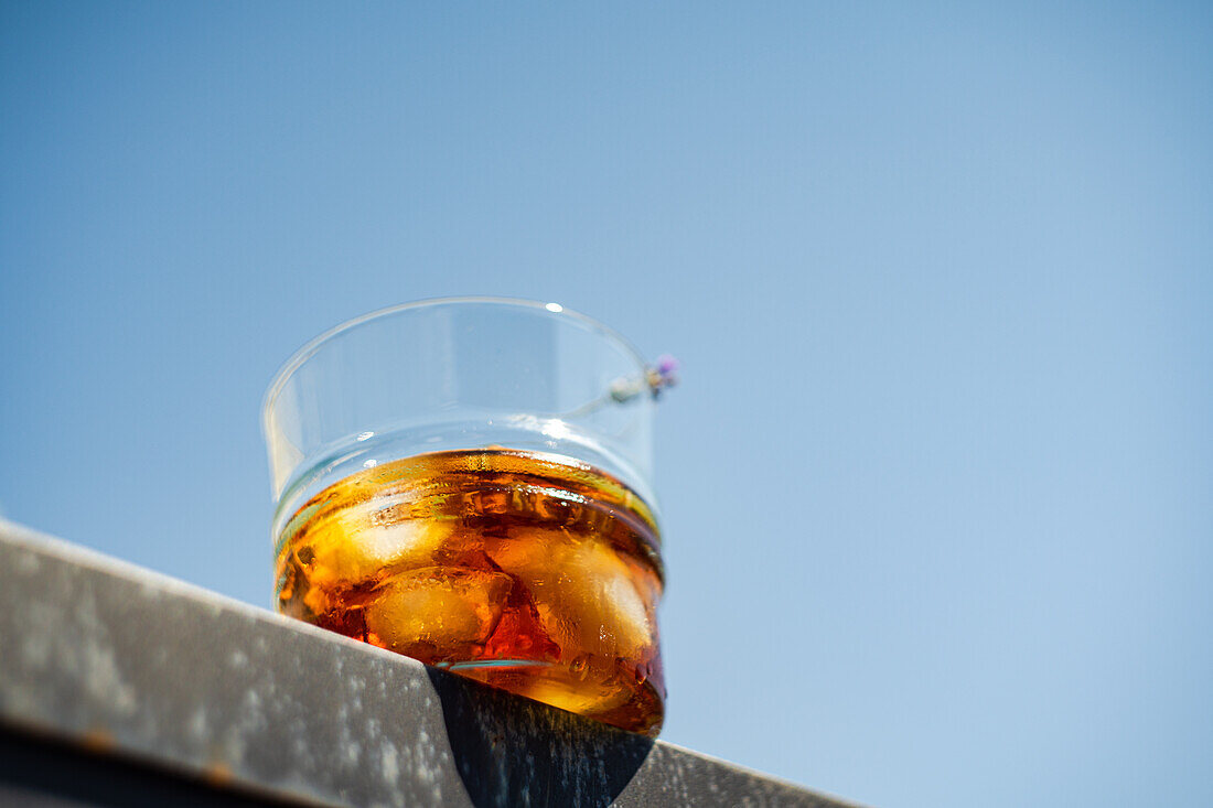 Ansicht von unten eines Whiskeyglases mit Eis, Orangenschale und Blume auf einer grauen Fläche vor blauem Himmel