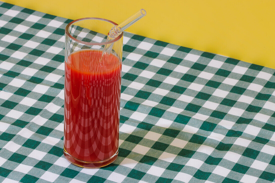 Transparentes Glas mit frischem Tomatensaft und Glasstrohhalm auf kariertem Tischtuch vor gelbem Hintergrund