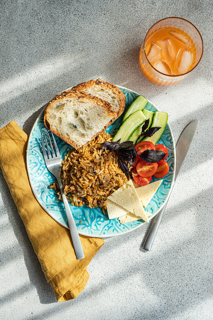 Draufsicht auf fünf Zutaten Mittagessen Set besteht aus hausgemachten Ziegenkäse, frische Gurken und Tomaten mit Blättern von Basilikum Kraut, zusätzlich zu gebratenem Reis mit Fleisch mit Scheiben von gesunden Sauerteig Brot serviert mit Glas Karotte-Apfel-Saft gegen grauen Hintergrund