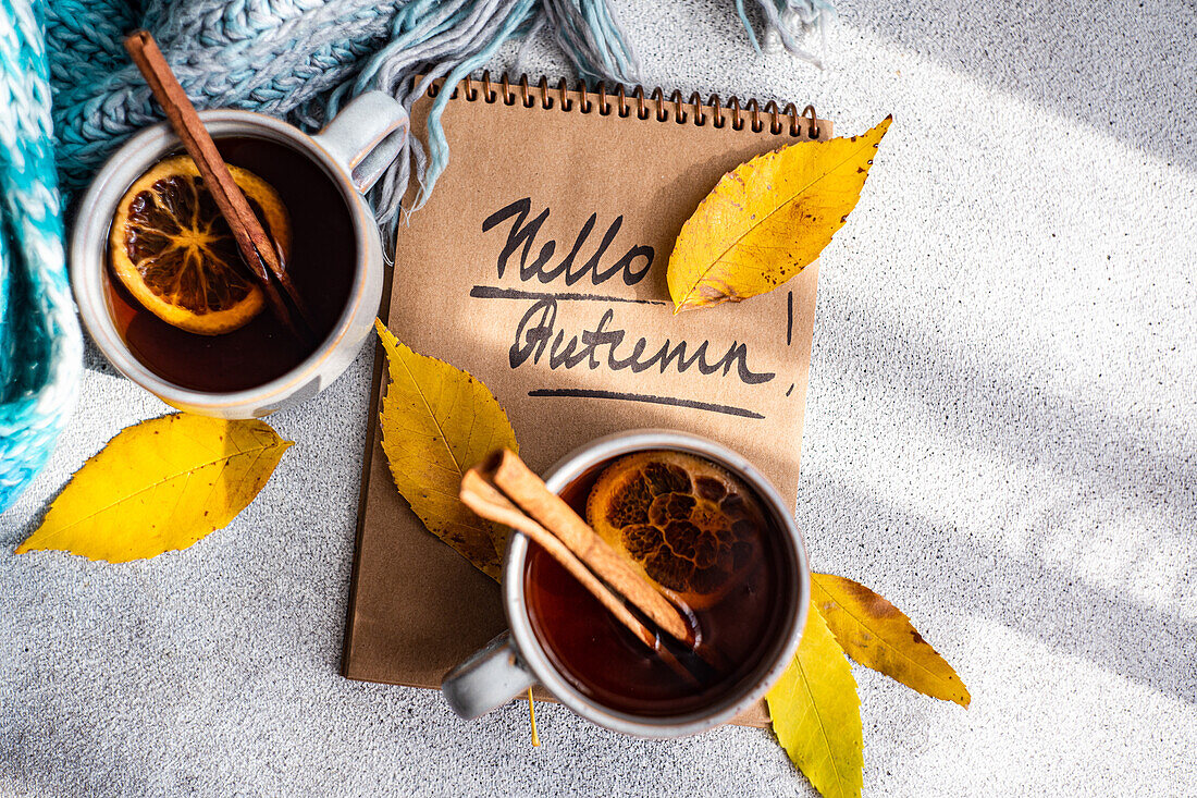 Top view of cups spiced tea with cinnamon sticks and dried orange slices, surrounded by vibrant yellow autumn leaves are juxtaposed against a notebook inscribed with "Hello Autumn!" on a textured gray background