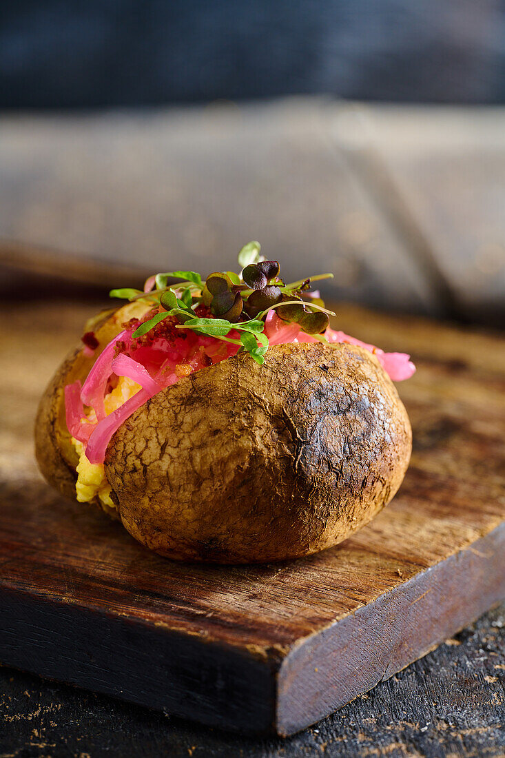Appetizing traditional Ecuadorian kumpir potato with stuffing and herbs placed on wooden cutting board against blurred background