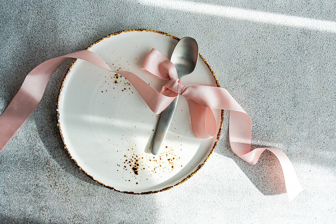 Ein leerer weißer Teller mit goldenem Rand, geschmückt mit einer zartrosa Schleife und einem silbernen Löffel, der elegante Schatten auf eine strukturierte Oberfläche wirft