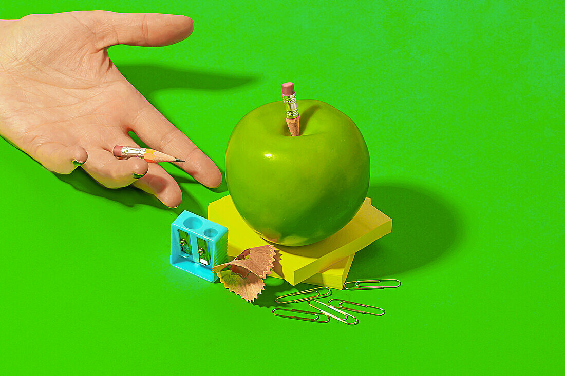 From above of crop hand of anonymous person demonstrating green apple with school supplies on bright color background